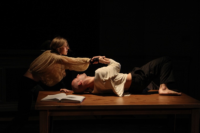 Chelsea Bonosky holds Adam Barruch's Wrist as he lays on his back on a wooden table. A book lays open in the foreground.